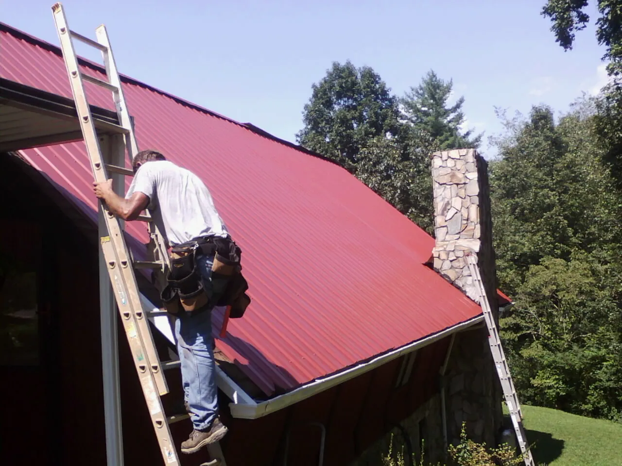 Roofer on a ladder beside a red metal roof and stone chimney — Soffit & Fascia Repair by Roofing Brothers in Earlysville